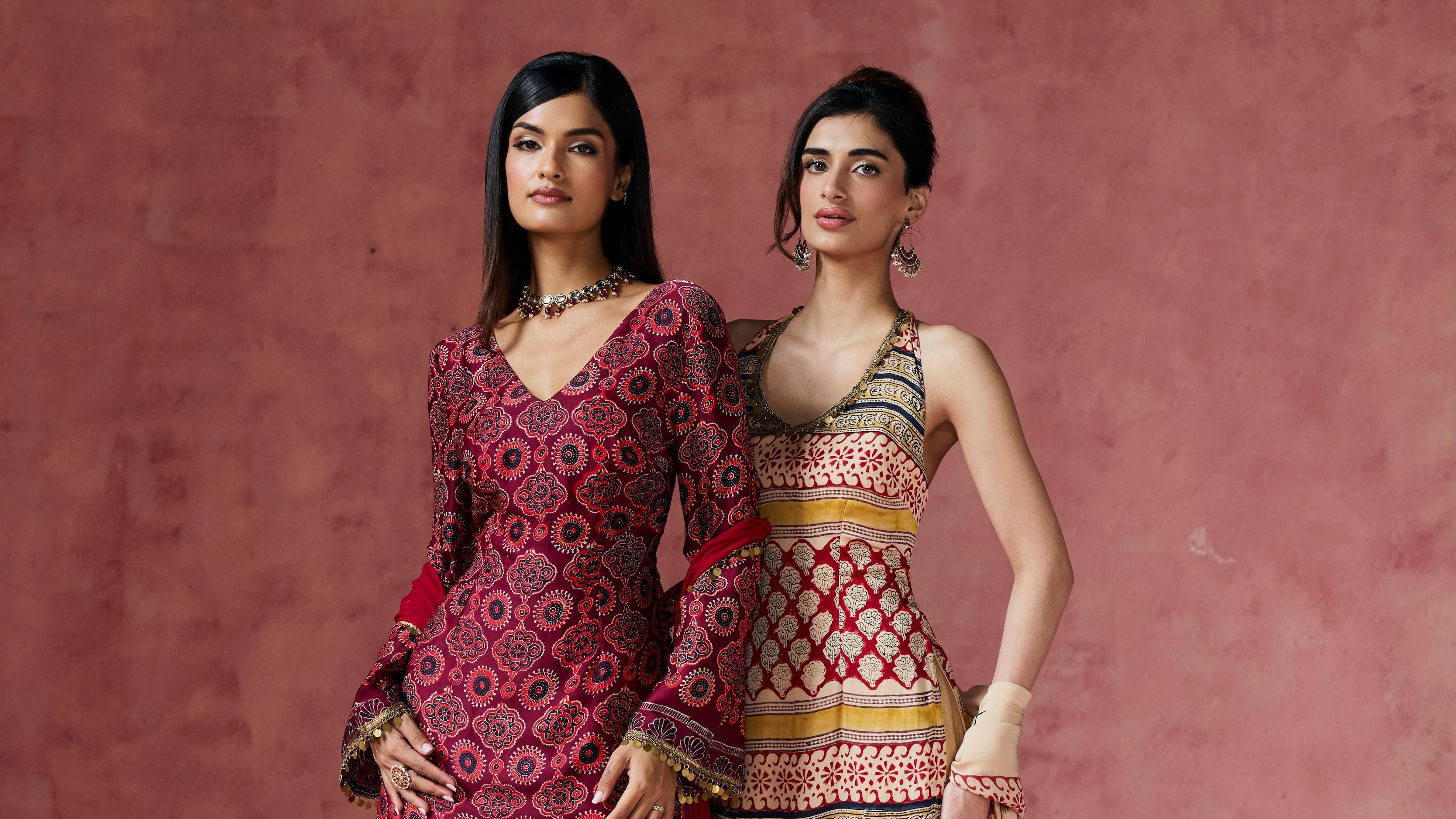 Two women modeling traditional South Asian salwar kameez and sharara suits against a textured dusty pink backdrop. The woman on the left wears a deep magenta patterned kameez with matching wide-leg shararas.